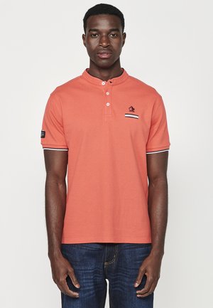 Īsām piedurkņu oranžs polo krekls ar henley tipa apkakli, baltiem akcentiem uz piedurknēm un mazu izšūtu logo uz krūšu kabatas.