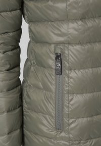 Gros plan d'une veste matelassée vert olive avec poche zippée verticale et tirette noire sur le devant.