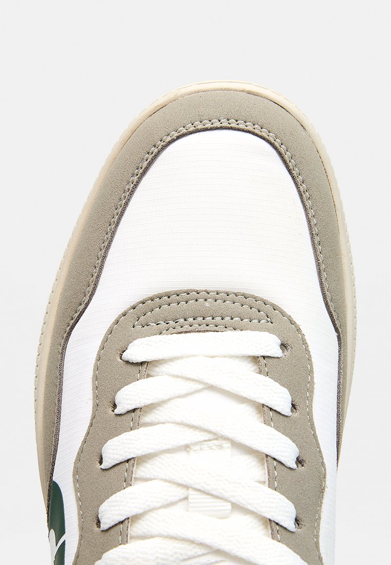 Zapatilla para hombre de lona blanca con detalles en ante gris. Presenta puntera redonda, cordones planos blancos y una suela de goma lisa. Detalle de marca en color verde.