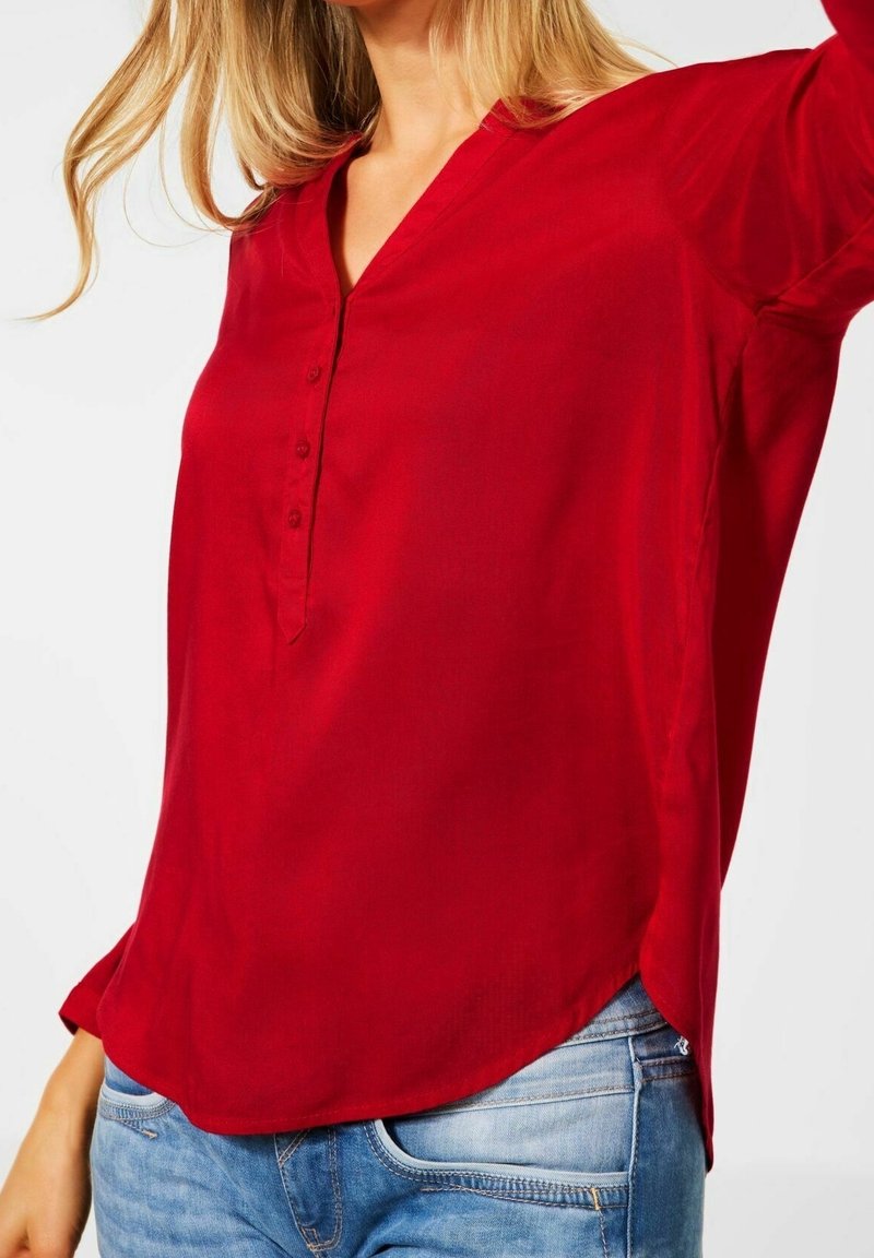 Blouse rouge à manches longues avec un décolleté en V et une petite patte de boutonnage. Tissu doux, ourlet légèrement courbé et coupe décontractée. Associée à un jean bleu.