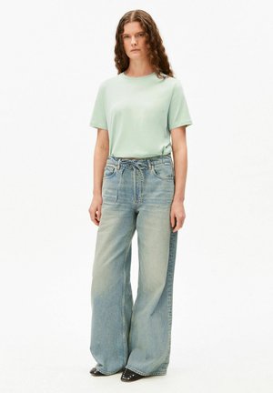 Vrouw met krullend haar, gekleed in een lichtgroen shirt met korte mouwen, een broek met wijde pijpen in een vervaagd blauw denim en zwarte schoenen met studs.