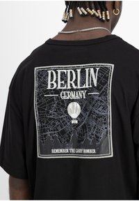 T-shirt noir avec un imprimé graphique représentant une carte de Berlin, "BERLIN ALLEMAGNE", avec une montgolfière et des silhouettes en bas.