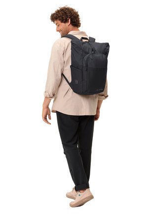 Sac à dos noir avec compartiments zippés, bretelles rembourrées et poches latérales en mesh, porté sur le dos, complété par une chemise beige et un pantalon sombre.
