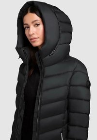 Kvinde iført en sort dunjakke med en oversized hætte, som er let åbnet, mod en ensfarvet lys grå baggrund.