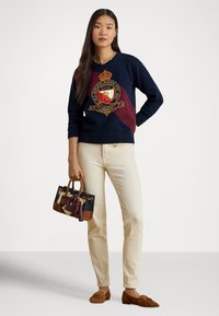 Μπλε ναυτικό και μπορντό πουλόβερ με λογότυπο, κρεμ παντελόνι και μια μικρή τσάντα με σχεδιαστικό μοτίβο. Καφέ μοκασίνια από σουέντ ολοκληρώνουν την εμφάνιση.