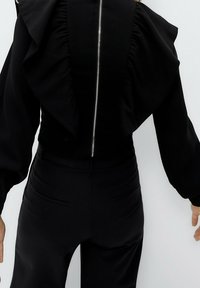 Černý top s řasením na ramenech, se zapínáním na zip na zadní straně. V kombinaci s elegantními černými kalhotami se zadními kapsami. Hladká textura.