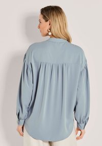 Frau mit blonden Haaren, die eine lockere, hellblaue Langarmbluse mit gerafftem Rücken und großen Perlenhängern trägt.