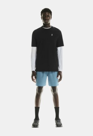 T-shirt noir à manches courtes par-dessus une chemise blanche à manches longues, associé à un short bleu clair et des chaussettes et chaussures de sport noires.