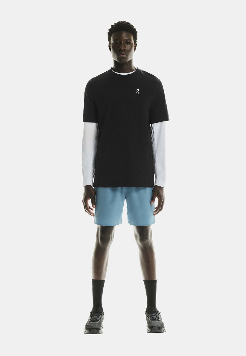 Camiseta negra de manga corta sobre una camisa blanca de manga larga, combinada con pantalones cortos azul claro y calcetines y zapatos deportivos negros.