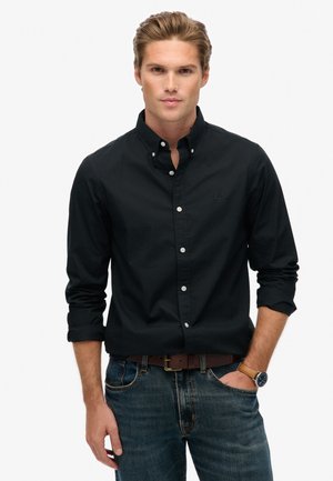 Camisa negra de botones con cuello, de mangas largas y botones blancos. Combinada con jeans azules y un cinturón marrón. Sin patrones o texturas notables.