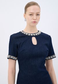 Tummansininen farkkukleid, jossa on lyhyet hihat, koristeltu kaula-aukko ja hihansuut, joissa on hopeisia kukkakuviota. Mekossa on myös avonainen kaula-aukko.