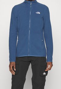 Niebieska bluza polarowa z wysokim kołnierzem, pełnym zamkiem błyskawicznym, kieszeniami po bokach oraz białym logo The North Face na lewej piersi. Gładka faktura.