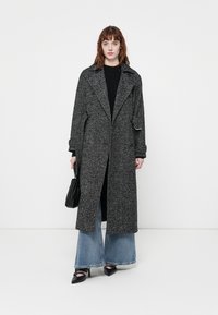 Manteau en chevrons noir et blanc avec un col cranté, des manches longues, des poignets larges et une fente sur le côté. Associé à un jean évasé et des chaussures noires.