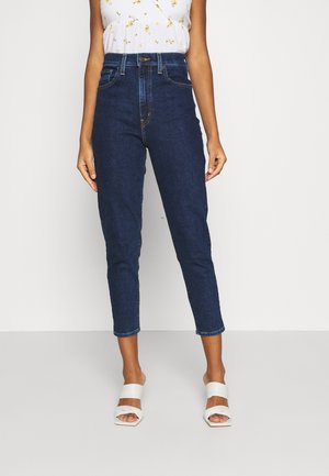 Vrouw draagt donkerblauwe jeans met hoge taille, witte mouwloze blouse met bloemenprint en witte open teen sandalen met hak, staand tegen een witte achtergrond.