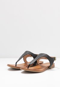 Sandalias de imitación de cuero negras con un diseño de tiras texturizadas y una suela plana color beige. Contienen un acabado de hebilla dorada en la correa del tobillo.