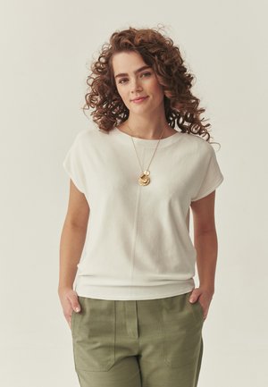 Young woman with curly hair wearing white short-sleeve top, green pants, and layered gold pendant necklace, hands in pockets, neutral background.