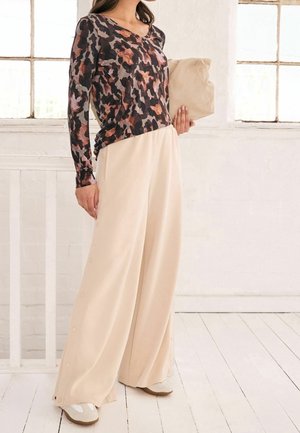 Une femme est debout à l'intérieur, portant un haut à fleurs à manches longues, un pantalon large beige avec des boutons latéraux, des chaussures blanches, et tenant une pochette beige.