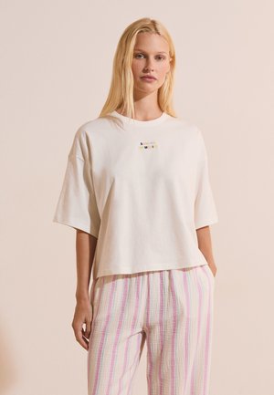 Kobieta z długimi blond włosami, ubrana w oversize białą koszulkę z napisem „SUMMER MEMORIES” oraz jasne, w paski spodnie, stojąca na jednolitym tle.