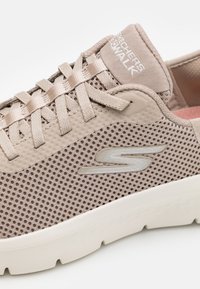 Tenis bej ușor, cu stabilitate din plasă, vârf rotund și latură texturată. Prezintă un logo și șireturi plate, combinate cu o talpă amortizată.