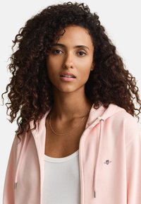 Junge Frau mit lockigem dunklem Haar, die einen hellrosa Kapuzenpullover mit Reißverschluss über einem weißen, gerippten Top trägt, blickt nach vorne vor weißem Hintergrund.