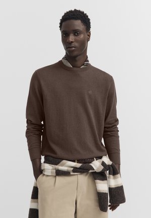 Junger Mann, der einen braunen Pullover über einem gestreiften Hemd trägt und einen schwarz-weißen Schal um beige Hosen gebunden hat, vor einem einfarbigen Hintergrund.