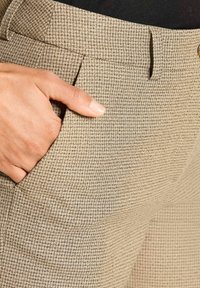Hand in de voorzak van beige geruite broek met plooien en riemlussen, gedragen met een zwart top.