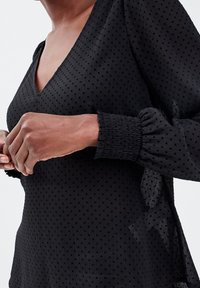 Nainen, jonka yllään on läpinäkyvä musta pallokuvioinen paita V-kaula-aukolla ja kuminauhakujilla, kädet ristissä vyötäisillä, vaaleaa yksinkertaista taustaa vasten.