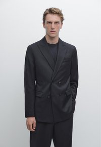 Blazer nero doppiopetto con taglio su misura, revers a punta e tasche laterali. Indossato sopra una maglietta nera, con una texture liscia.