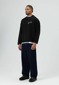 Zwarte trui met borstzak en wit schriftontwerp, gecombineerd met losse, donkerblauwe jeans en stevige zwarte schoenen.