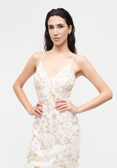 Γυναίκα με μακριά σκουρόχρωμα μαλλιά, που φοράει λευκό φόρεμα με λεπτές τιράντες και κεντήματα με λουλούδια, ποζάρει με τα χέρια στη μέση μπροστά σε γκρι φόντο.