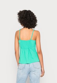 Top sem mangas, ligeiramente evasé, em turquesa brilhante, com parte de trás franzida, apresentando tiras finas e tecido texturizado, combinado com jeans azul claro.