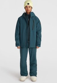 Veste de ski teal avec capuche et fermeture éclair, accompagnée d'un pantalon assorti. Les caractéristiques incluent des coutures prononcées et des accents de logo. Matériau chaud et isolé.