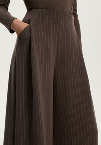 Bruine, wijde broek met verticale streepjes, gemaakt van een zachte, rekbare stof. Beschikt over een zijzak en geplooid ontwerp.