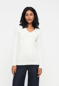 Maglione bianco a maniche lunghe con scollo a V, dal design aderente e con il logo "Guess" testurizzato sul davanti, abbinato a jeans larghi scuri.