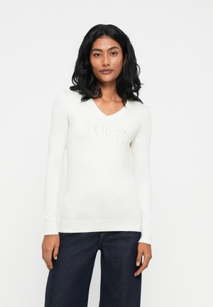 Maglione bianco a maniche lunghe con scollo a V, dal design aderente e con il logo "Guess" testurizzato sul davanti, abbinato a jeans larghi scuri.