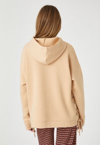 Beżowy hoodie z dużym kapturem, opuszczonymi ramionami i sznurkami po bokach. Miękka tkanina i luźny krój, zestawiony z wzorzystymi legginsami.