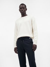 Homme portant un pull en tricot blanc et un pantalon noir à cordon, debout devant un fond blanc uni.