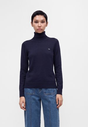 Femme portant un pull à col roulé bleu marine foncé et un jean bleu, debout devant un fond clair uni, face à elle.