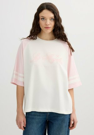 Mujer con cabello castaño ondulado que lleva una camiseta blanca holgada con mangas rosas y franjas rosa claro cerca de los codos, combinada con vaqueros azules.