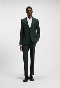 Dunkelgrüner maßgeschneiderter Anzug mit slim-fit Hose, einreihigem Blazer und einem weißen Hemd darunter. Schwarze Schuhe runden den Look ab.
