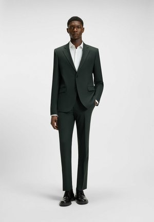 Dark green tailored suit with slim-fit trousers, single-button blazer, and a white shirt underneath. Black shoes complete the look.