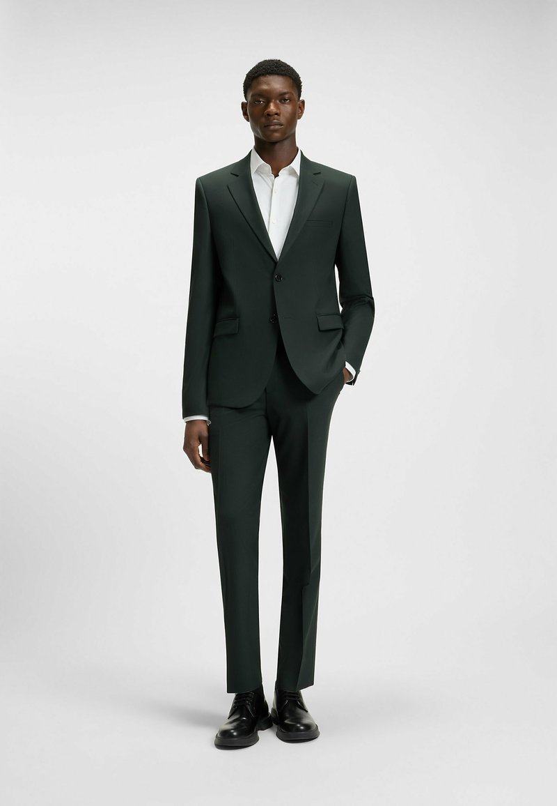 Dunkelgrüner maßgeschneiderter Anzug mit slim-fit Hose, einreihigem Blazer und einem weißen Hemd darunter. Schwarze Schuhe runden den Look ab.