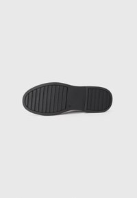 Suela de zapato de goma negra con surcos texturizados, con estrías paralelas para el agarre. Superficie suave cerca del talón con marcas sutiles.