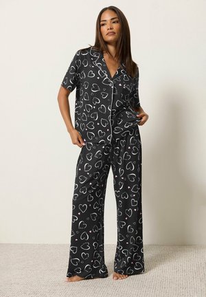 Femme portant un ensemble de pyjama noir avec des motifs de cœurs blancs et roses, debout pieds nus contre un mur clair uni et un sol recouvert de moquette.
