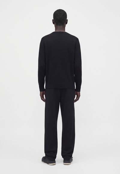 Homme portant une chemise noire à manches longues, un pantalon noir ample et des chaussures noires, debout de dos devant un fond gris clair uni.