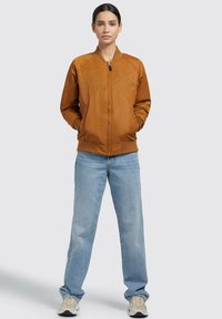 Giacca bomber marrone con ricamo sul petto, zipper frontale, polsini e orlo a costine, abbinata a jeans di denim azzurri e sneakers.