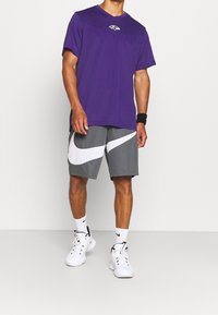 Fialová košile s krátkým rukávem se znakem, v kombinaci se šedými šortkami se velkým bílým logo Nike swoosh. Doplněno bílými teniskami Nike a bílými ponožkami.