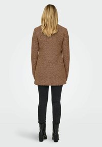 Abrigo marrón texturizado con mangas largas, corte recto y abertura en la parte trasera. Combinado con jeans skinny negros y botas negras de tacón.
