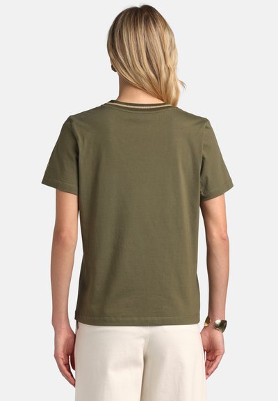 Femme aux cheveux longs et blonds portant un t-shirt manches courtes vert olive et un pantalon blanc, debout dos à la caméra.