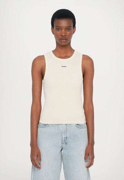 Light beige ribbed tank top with round neckline and logo detail. Paired with light blue loose-fitting denim jeans. Simple, fitted design.
